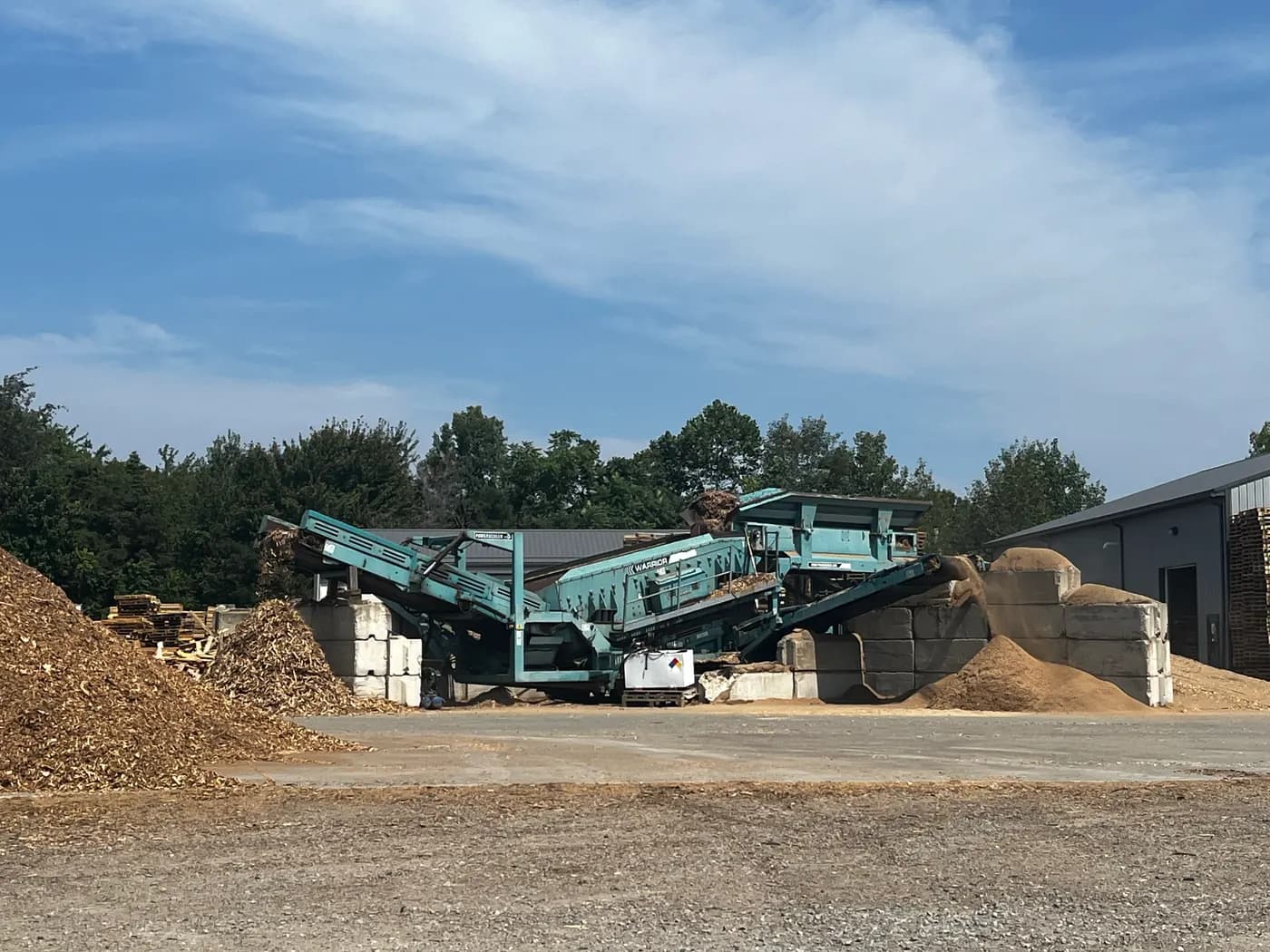 Wood-recycling yard with a green grinder and mounds of mulch under a blue sky, the ESG-aligned end-of-life wood stream behind remanufactured pallets