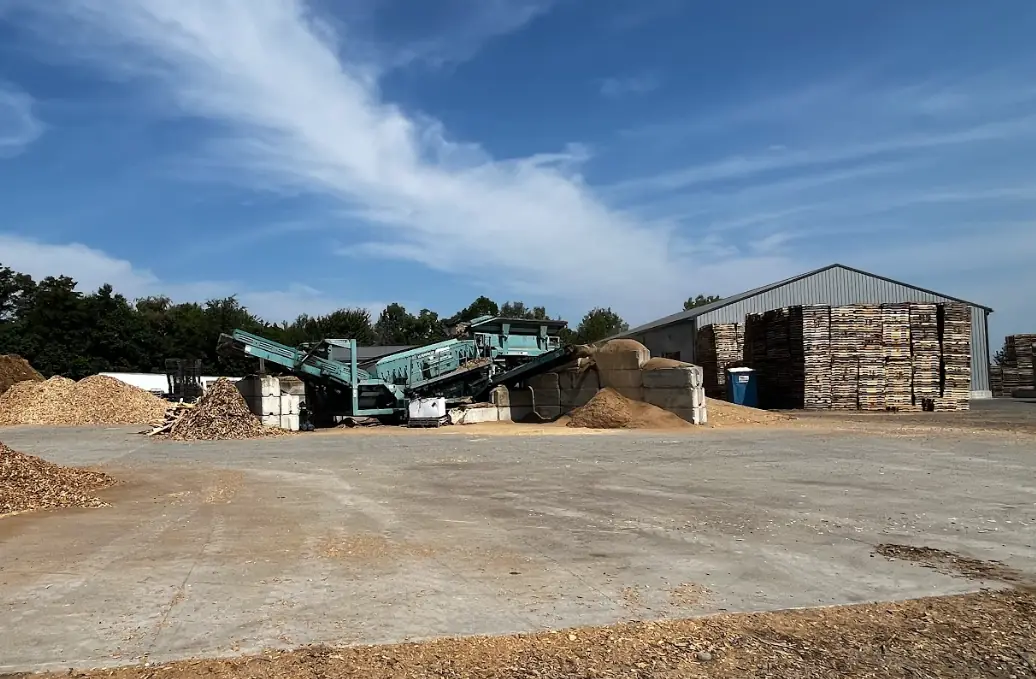 ePallet Sugarcreek recycling yard under a bright blue sky — a green wood grinder, sawdust and mulch piles, and a storage barn stacked with pallets